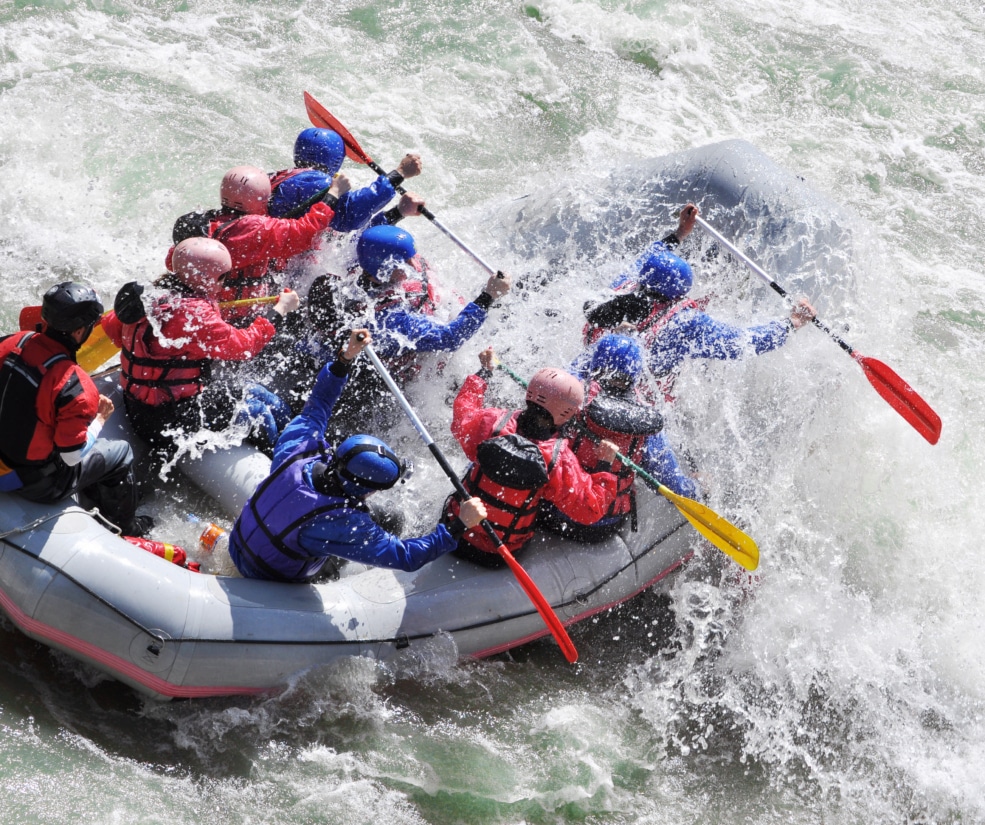 A large group of rafters in red and blue gear brace paddles as they navigate rough whitewater in a gray inflatable raft.