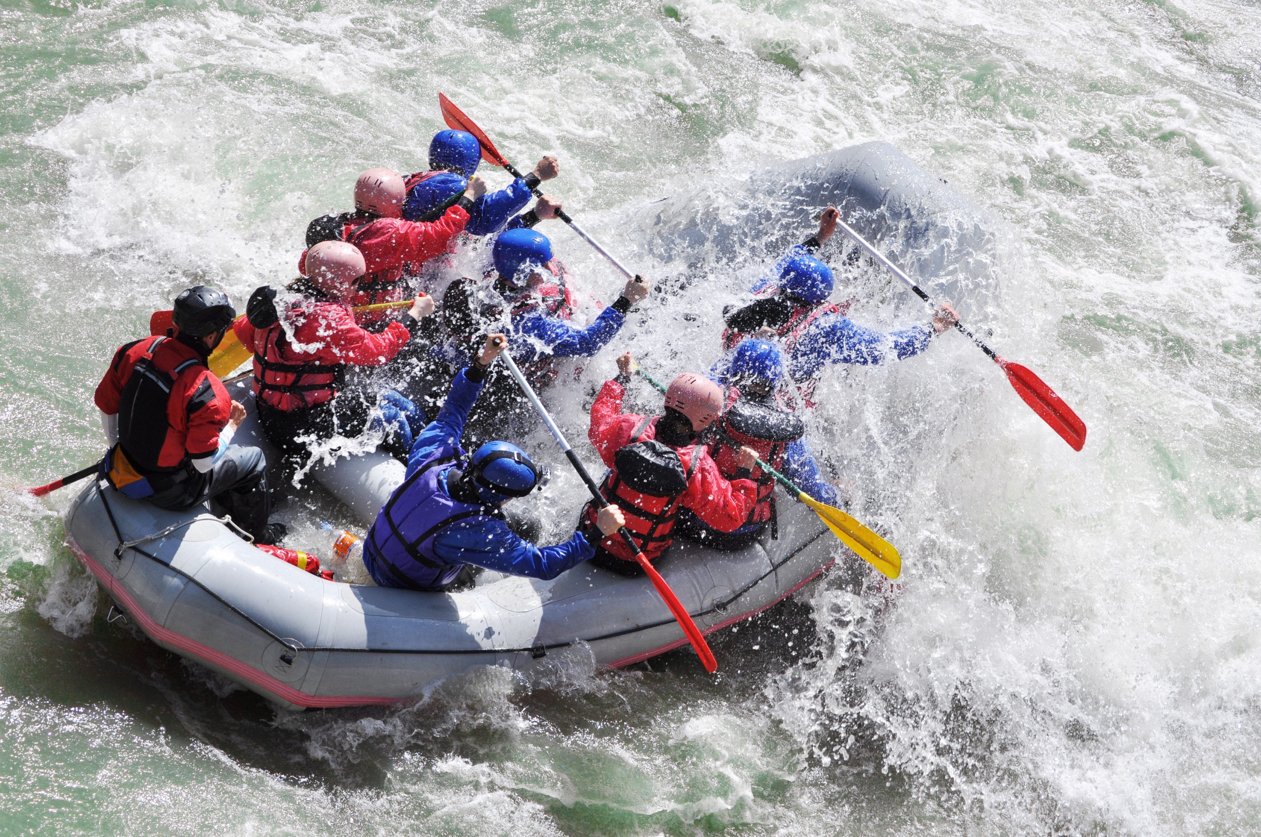 A large group of rafters in red and blue gear brace paddles as they navigate rough whitewater in a gray inflatable raft.