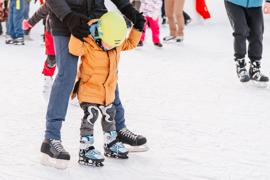 Child in an orange jacket and yellow helmet being assisted on the ice by an adult, with other skaters in the background on an ice rink.