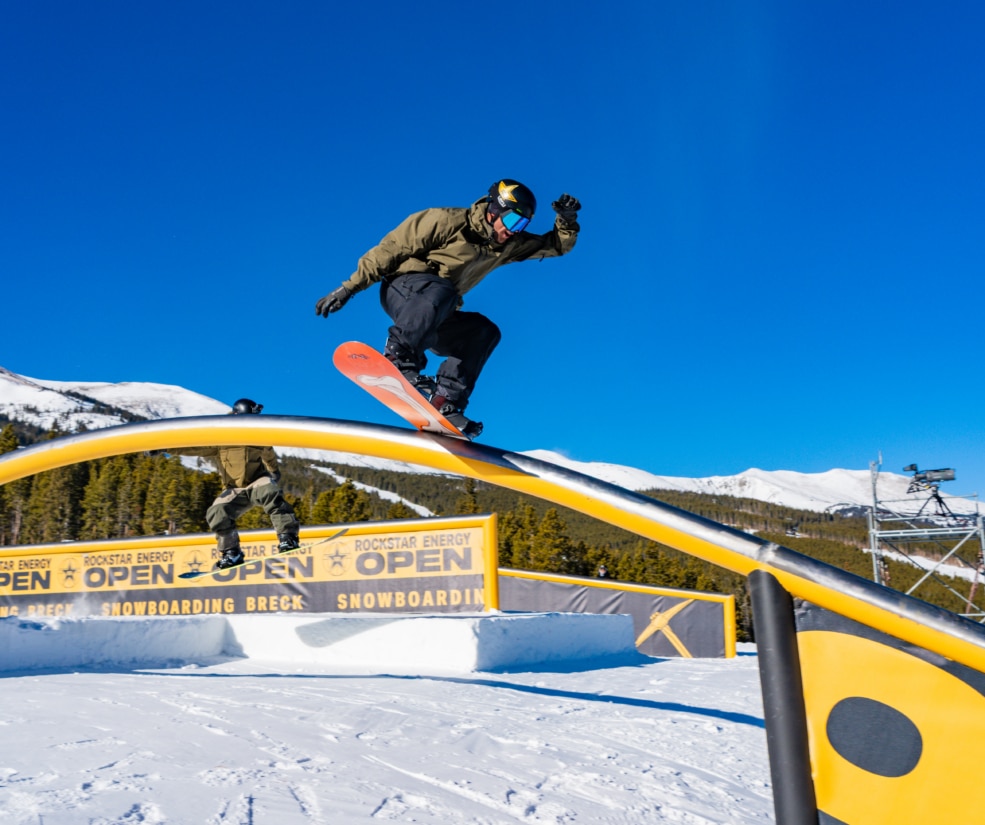 Snowboarder in a tan jacket and black pants jumping over a bright yellow rail against a clear blue sky.