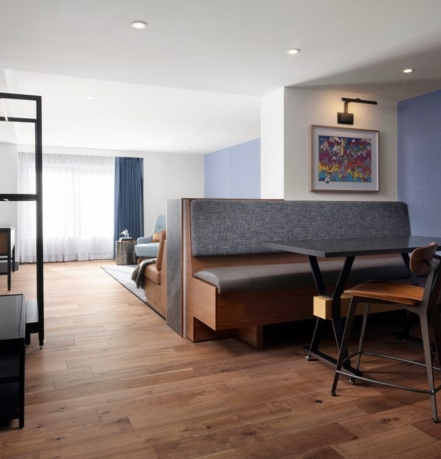 Open-plan living area with a booth-style dining table, wood chairs, and a blue accent wall beneath warm lighting. The space is bright from a large window with sheer curtains.
