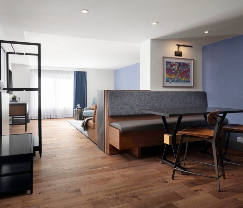 Open-plan living area with a booth-style dining table, wood chairs, and a blue accent wall beneath warm lighting. The space is bright from a large window with sheer curtains.