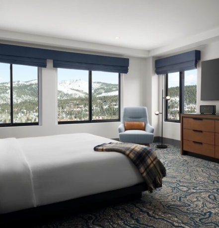 Hotel bedroom with a king bed, blue accents, and a mountainside view through large windows moldings.