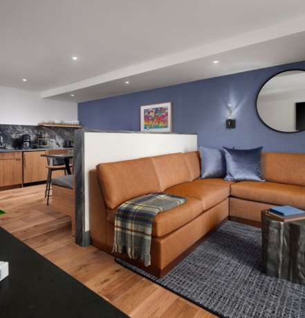 Open-plan living room with an orange leather sectional, blue accent wall, round wall mirror, and a dark metallic cube coffee table on a gray rug.