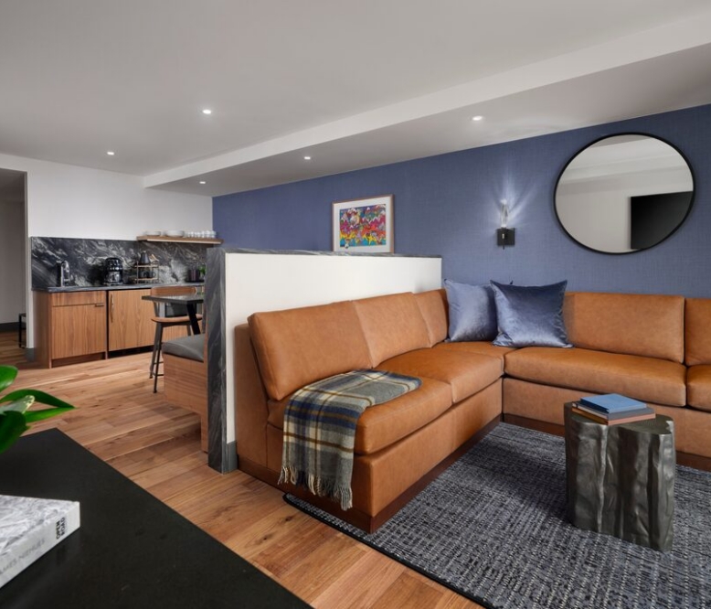Open-plan living room with an orange leather sectional, blue accent wall, round wall mirror, and a dark metallic cube coffee table on a gray rug.