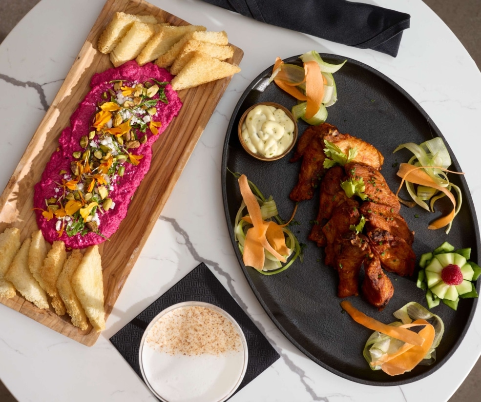 Plated appetizer spread from The Carter inside Hotel Alpenrock featuring crispy chicken wings with herb aioli and pickled vegetables alongside vibrant beet hummus topped with pistachios and edible flowers, served with toasted bread.