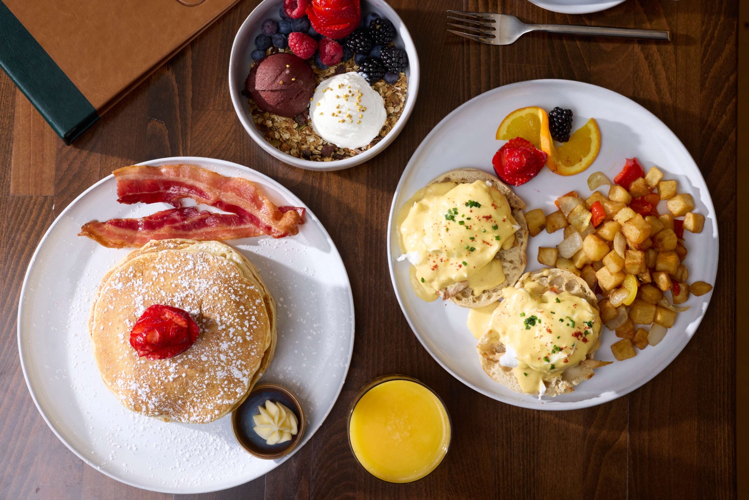 Breakfast spread from The Carter inside Hotel Alpenrock featuring pancakes with bacon, eggs Benedict with crispy potatoes, fresh fruit and granola, and a glass of orange juice.