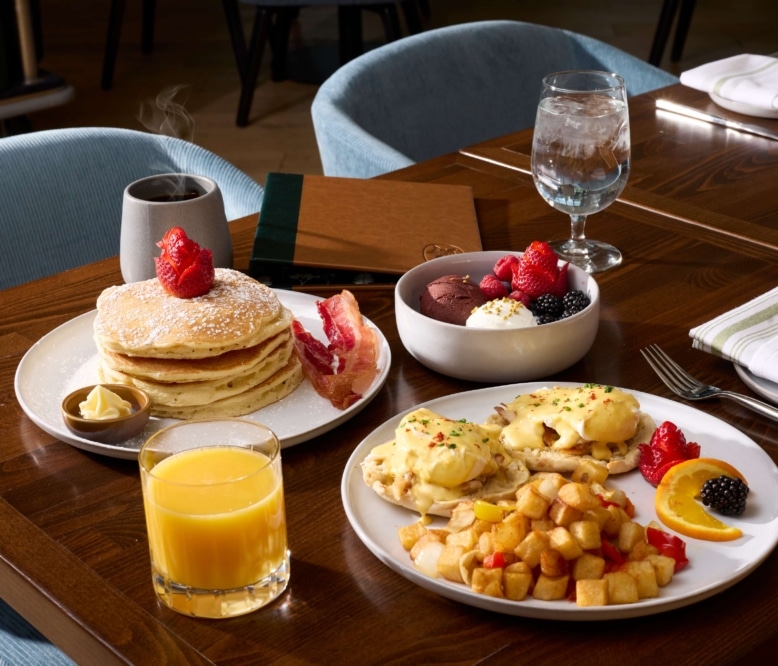 Breakfast spread from The Carter inside Hotel Alpenrock featuring pancakes with bacon, eggs Benedict with crispy potatoes, fresh fruit and granola, and a glass of orange juice.