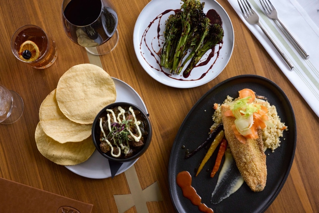 Assorted plated dishes from The Carter inside Hotel Alpenrock, including a breaded fish entrée over couscous with pickled vegetables, roasted broccolini drizzled with balsamic glaze, a savory dip served with crisp flatbread, and paired cocktails and red wine on a wooden table.