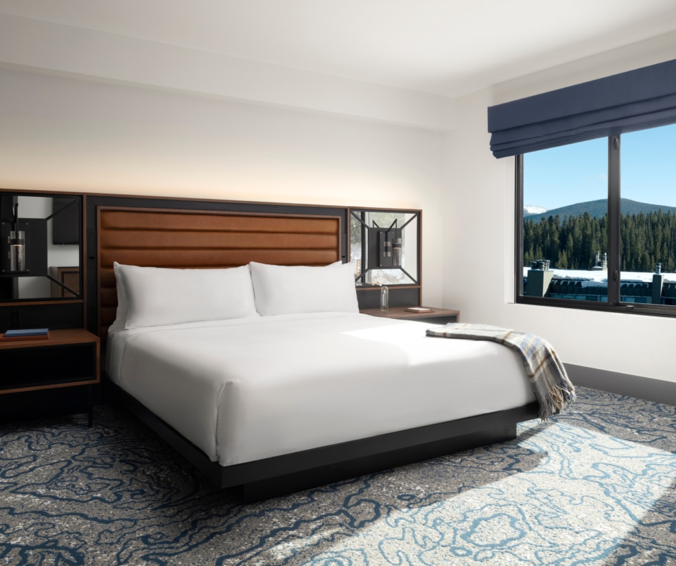King bedroom with plush white bedding, wood headboard, and mountain view window at Hotel Alpenrock.