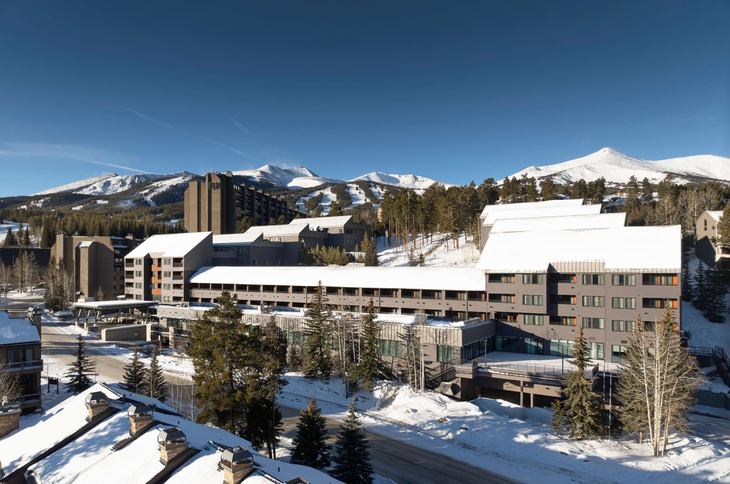 Daytime aerial view of Hotel Alpenrock with snow-covered rooftops, surrounding pine trees, and Breckenridge ski slopes in the background.