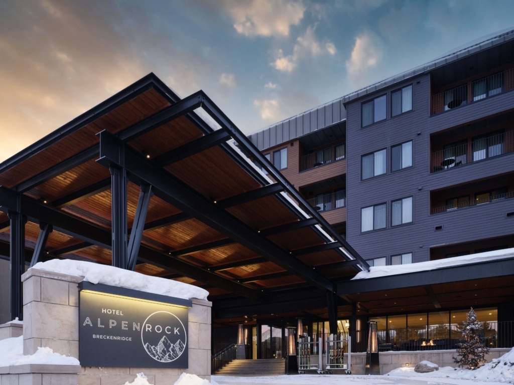 Dusk exterior view of Hotel Alpenrock in Breckenridge, featuring the illuminated entrance sign, modern covered porte cochere, and snow lined entry under a colorful evening sky.