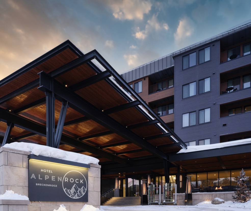Dusk exterior view of Hotel Alpenrock in Breckenridge, featuring the illuminated entrance sign, modern covered porte cochere, and snow lined entry under a colorful evening sky.