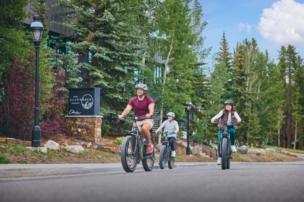 Guests riding electric bikes along the road at Hotel Alpenrock, surrounded by trees and mountain scenery.