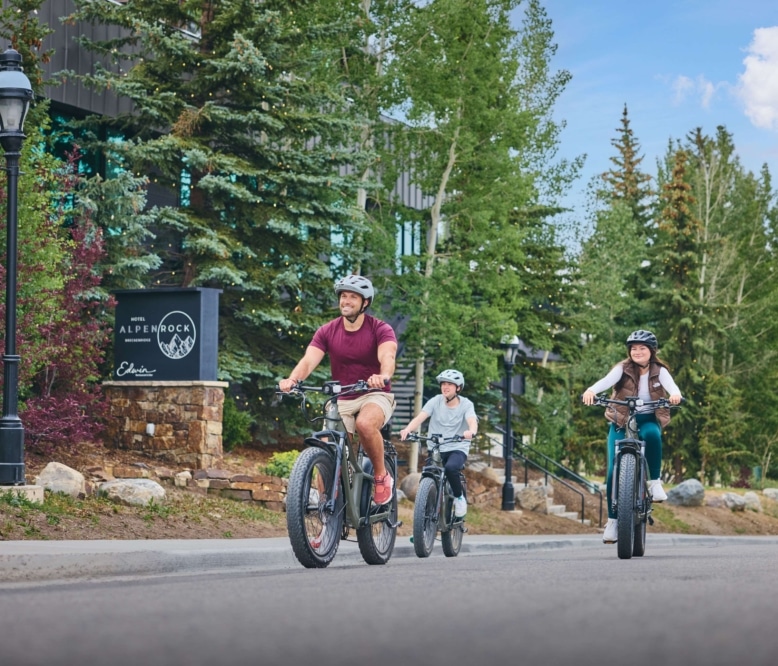 Guests riding electric bikes along the road at Hotel Alpenrock, surrounded by trees and mountain scenery.