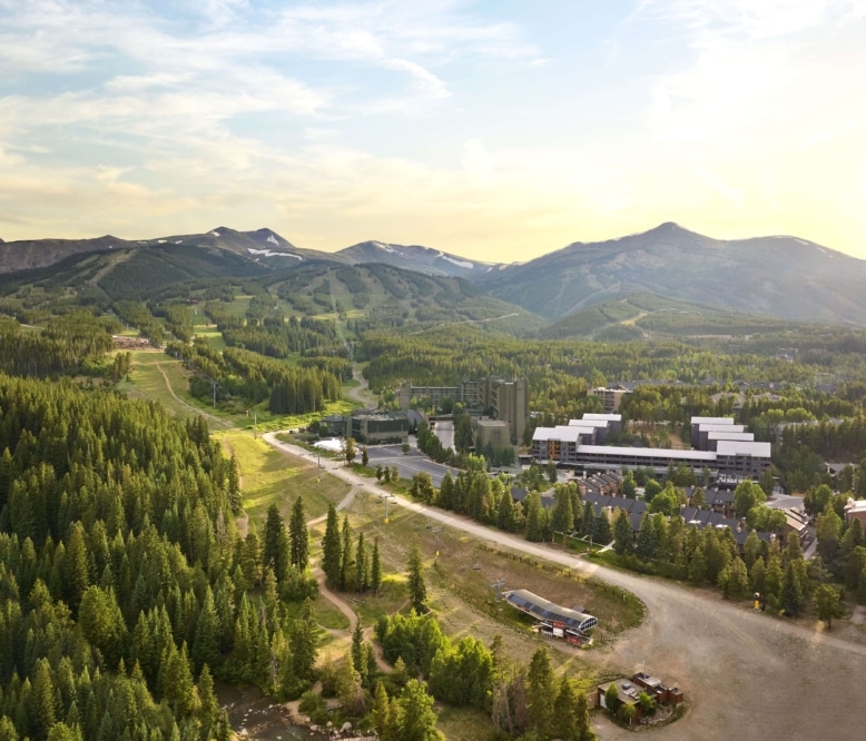 Aerial summer view of Hotel Alpenrock surrounded by lush pine forest, ski trails, and mountain peaks under warm evening light.