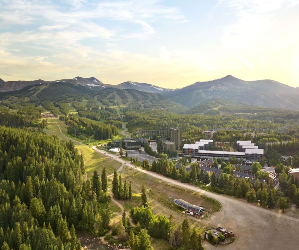 Aerial summer view of Hotel Alpenrock surrounded by lush pine forest, ski trails, and mountain peaks under warm evening light.