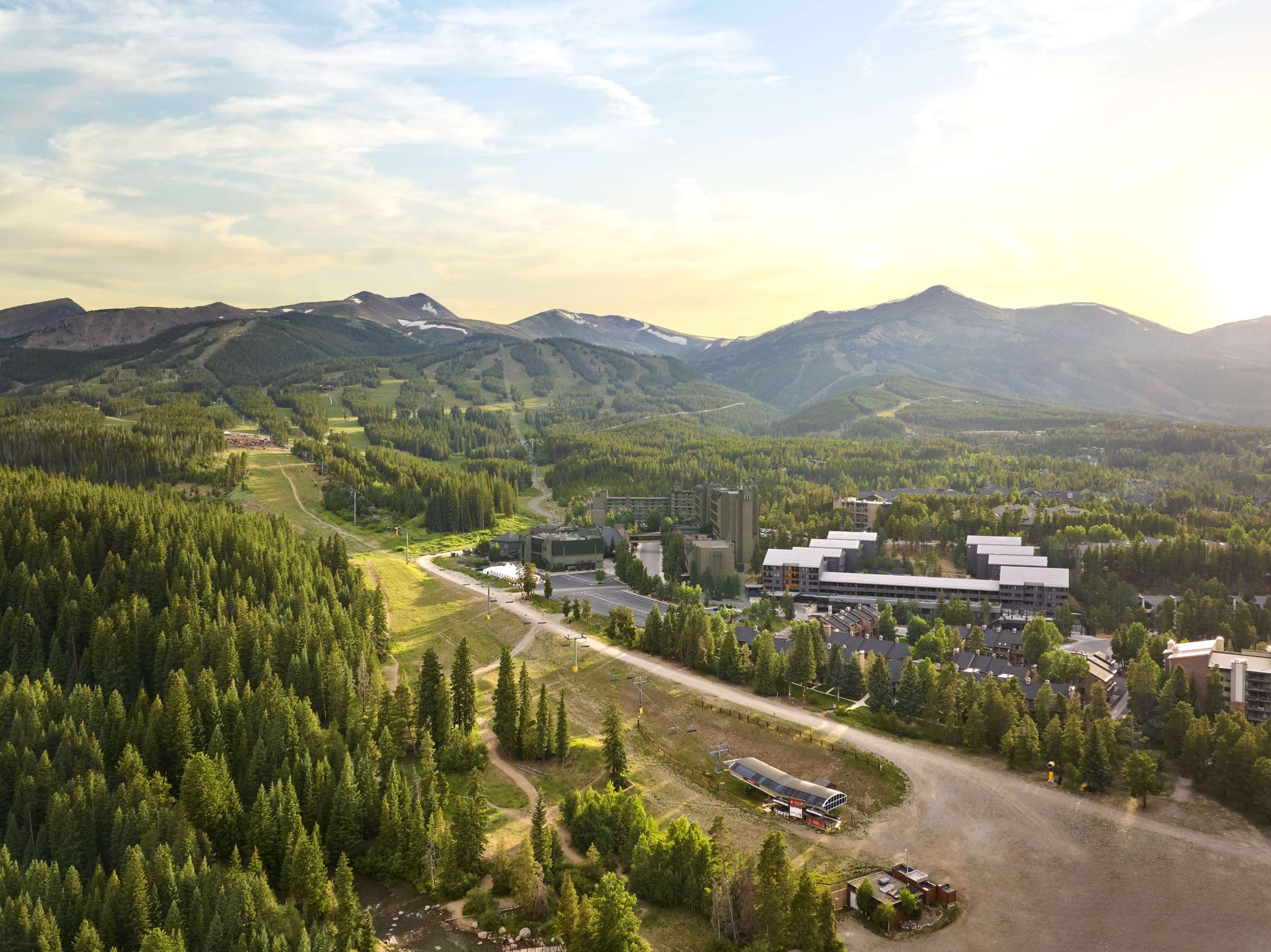 Aerial summer view of Hotel Alpenrock surrounded by lush pine forest, ski trails, and mountain peaks under warm evening light.