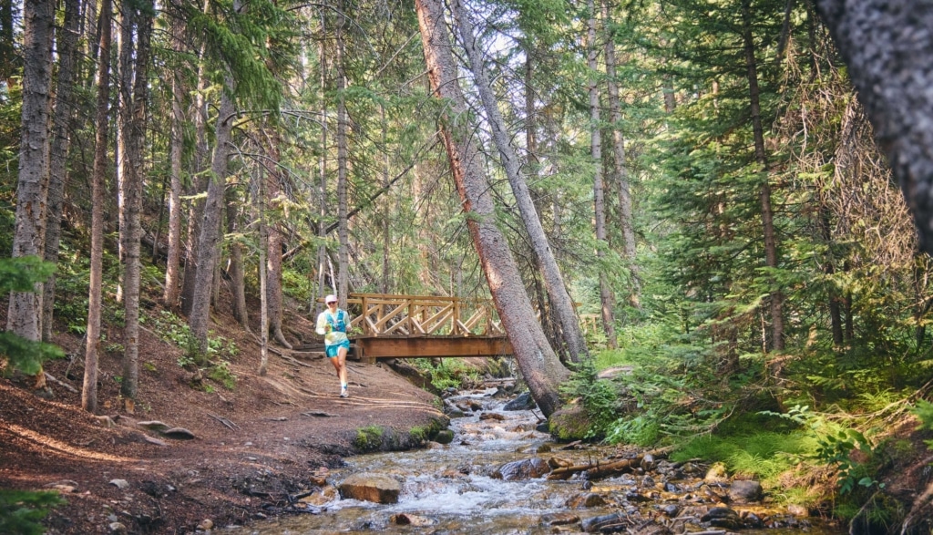 Trail runner moving along a forest path beside a creek and wooden bridge near Hotel Alpenrock.