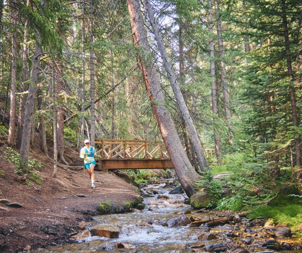 Trail runner moving along a forest path beside a creek and wooden bridge near Hotel Alpenrock.