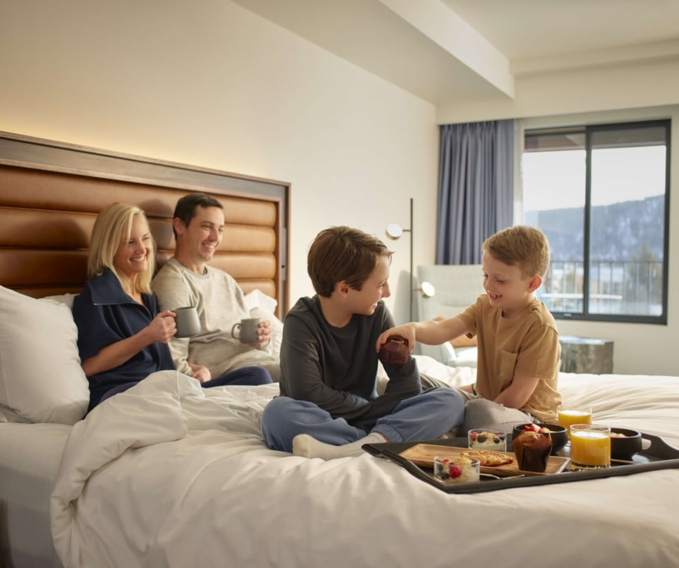 Family enjoying breakfast together on a bed inside a Hotel Alpenrock guest room.