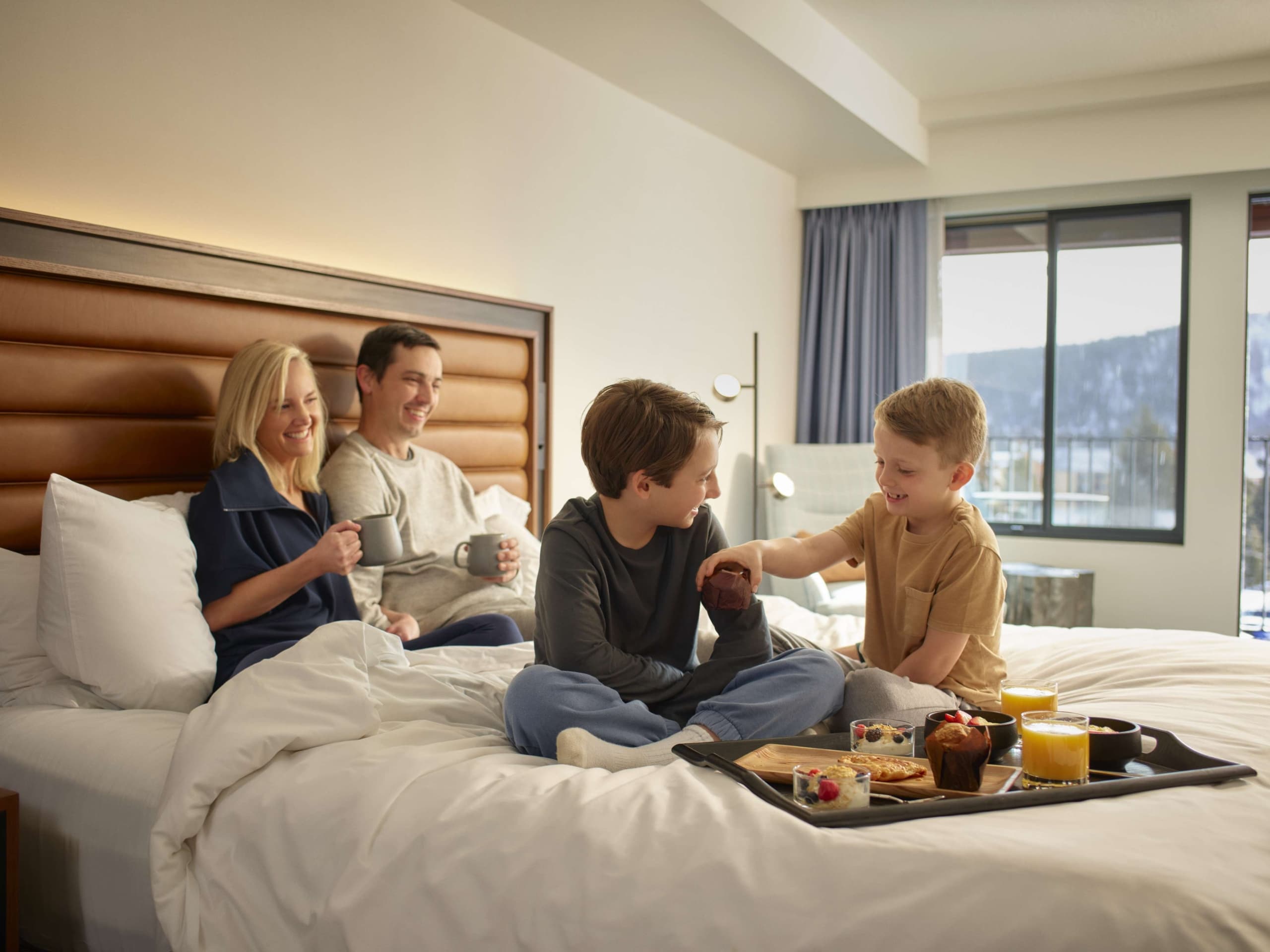 Family enjoying breakfast together on a bed inside a Hotel Alpenrock guest room.