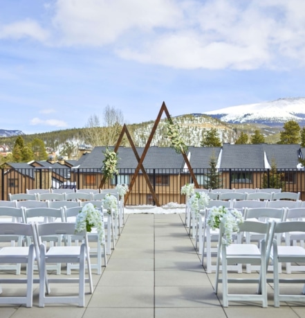 Outdoor wedding setup at Hotel Alpenrock with white chairs, floral-adorned triangular wooden arch, and panoramic snowy mountain views.