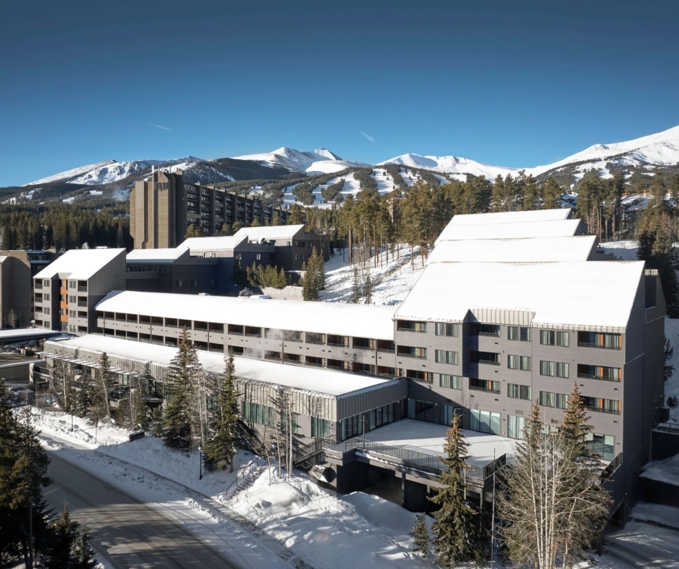 Daytime aerial view of Hotel Alpenrock with snow-covered buildings, surrounding pine forest, and Breckenridge ski slopes under a clear blue sky.