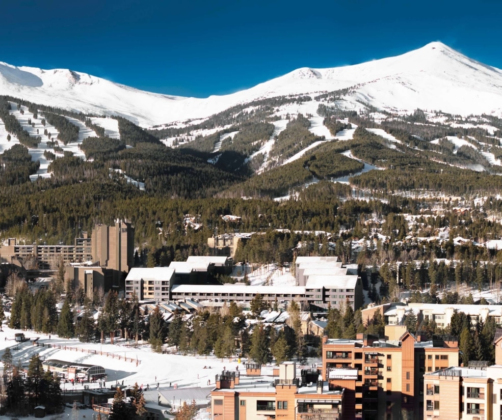 Panoramic winter view of Hotel Alpenrock in Breckenridge, with snow covered ski slopes, forested mountains, and surrounding village buildings under a clear blue sky.