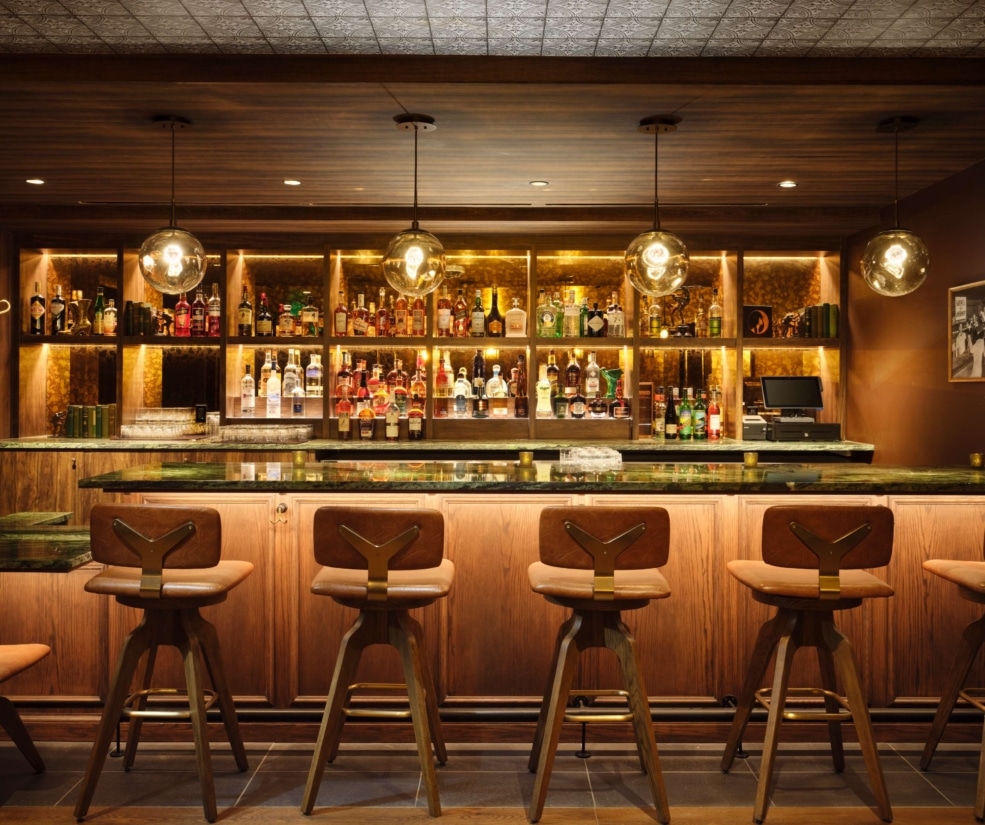 Warm speakeasy bar interior at The Edwin inside Hotel Alpenrock with backlit liquor shelves and wood bar seating.
