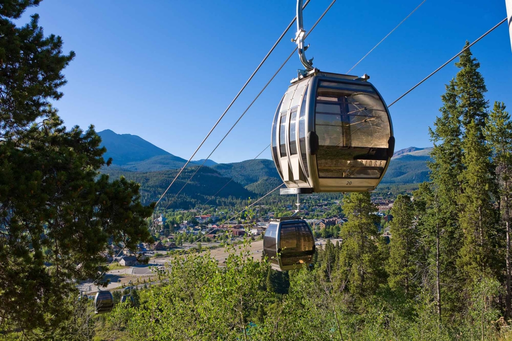 Cable car gondola suspended by cables over a forest with a mountain range and town below under a clear blue sky.
