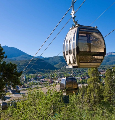 Cable car gondola suspended by cables over a forest with a mountain range and town below under a clear blue sky.
