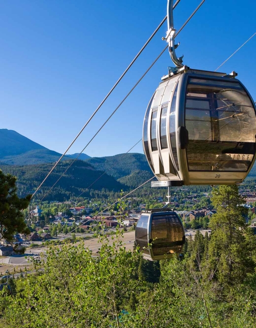 Cable car gondola suspended by cables over a forest with a mountain range and town below under a clear blue sky.
