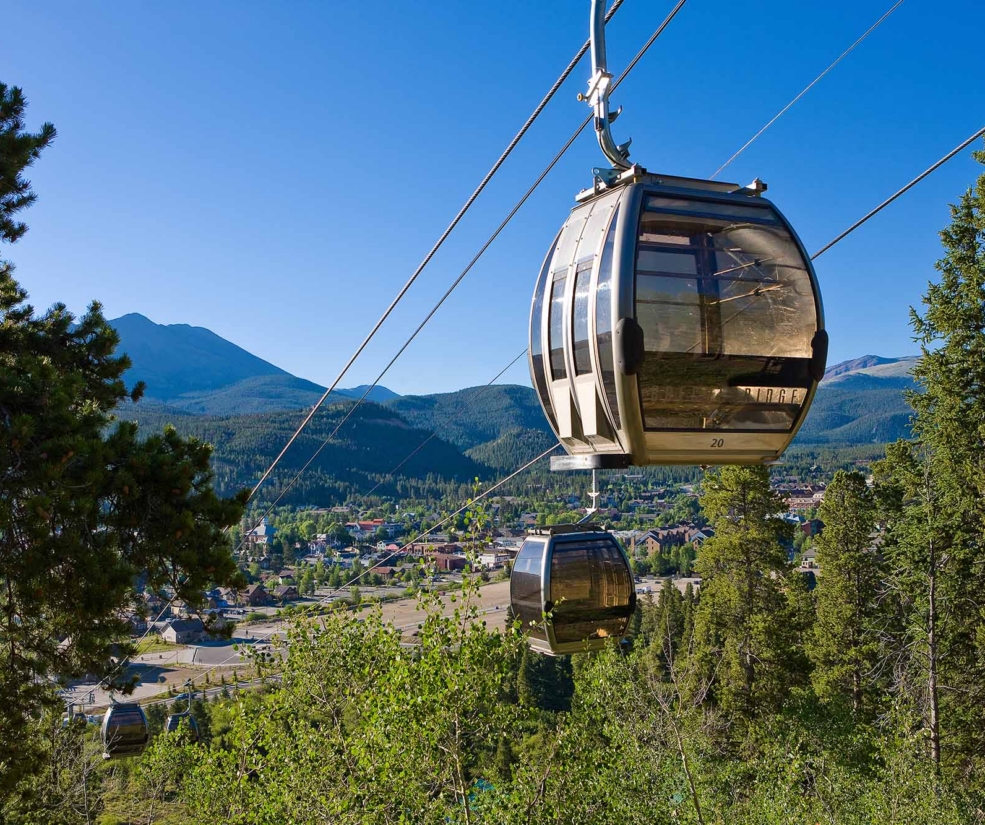 Cable car gondola suspended by cables over a forest with a mountain range and town below under a clear blue sky.