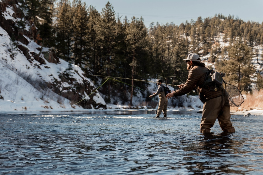 Two anglers wading in a snow-dusted river, casting lines in a forested, wintry landscape.