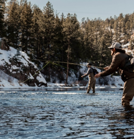 Two anglers wading in a snow-dusted river, casting lines in a forested, wintry landscape.