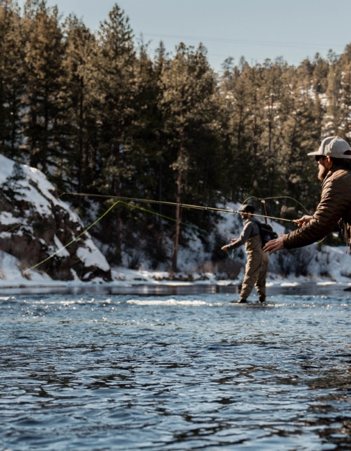 Two anglers wading in a snow-dusted river, casting lines in a forested, wintry landscape.