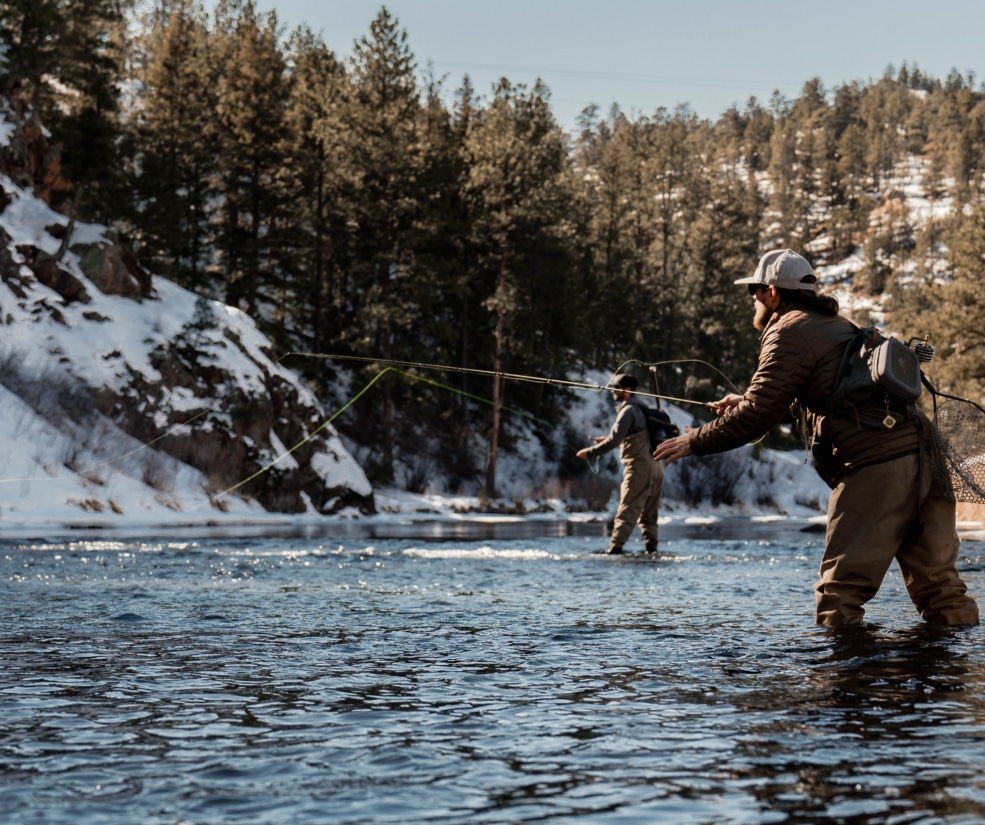 Two anglers wading in a snow-dusted river, casting lines in a forested, wintry landscape.