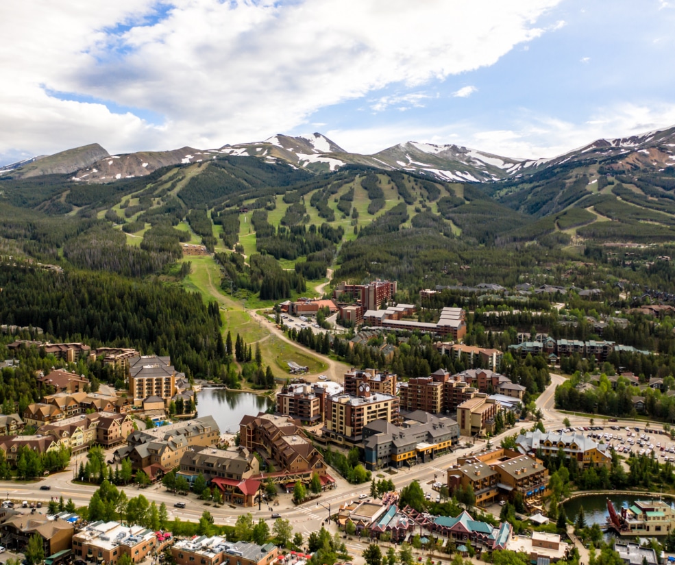 Aerial view of a mountain town with ski slopes, green hills, and clustered buildings below a cloudy sky.