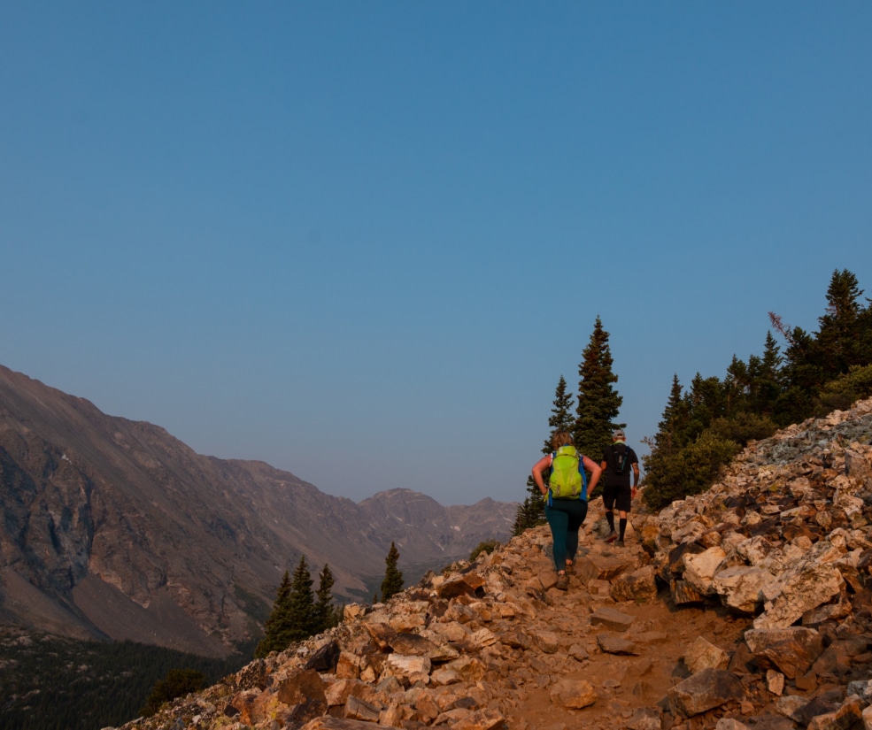 Two hikers with backpacks hike up a rocky mountain trail under a clear blue sky, with pine trees on the slope to the right.