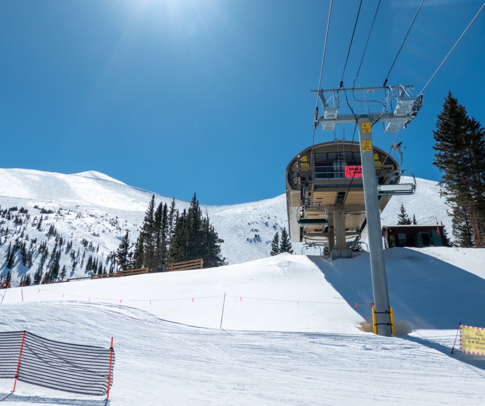 Ski lift rising from a snowy slope under a bright blue sky, with evergreen trees on the hillside nearby.
