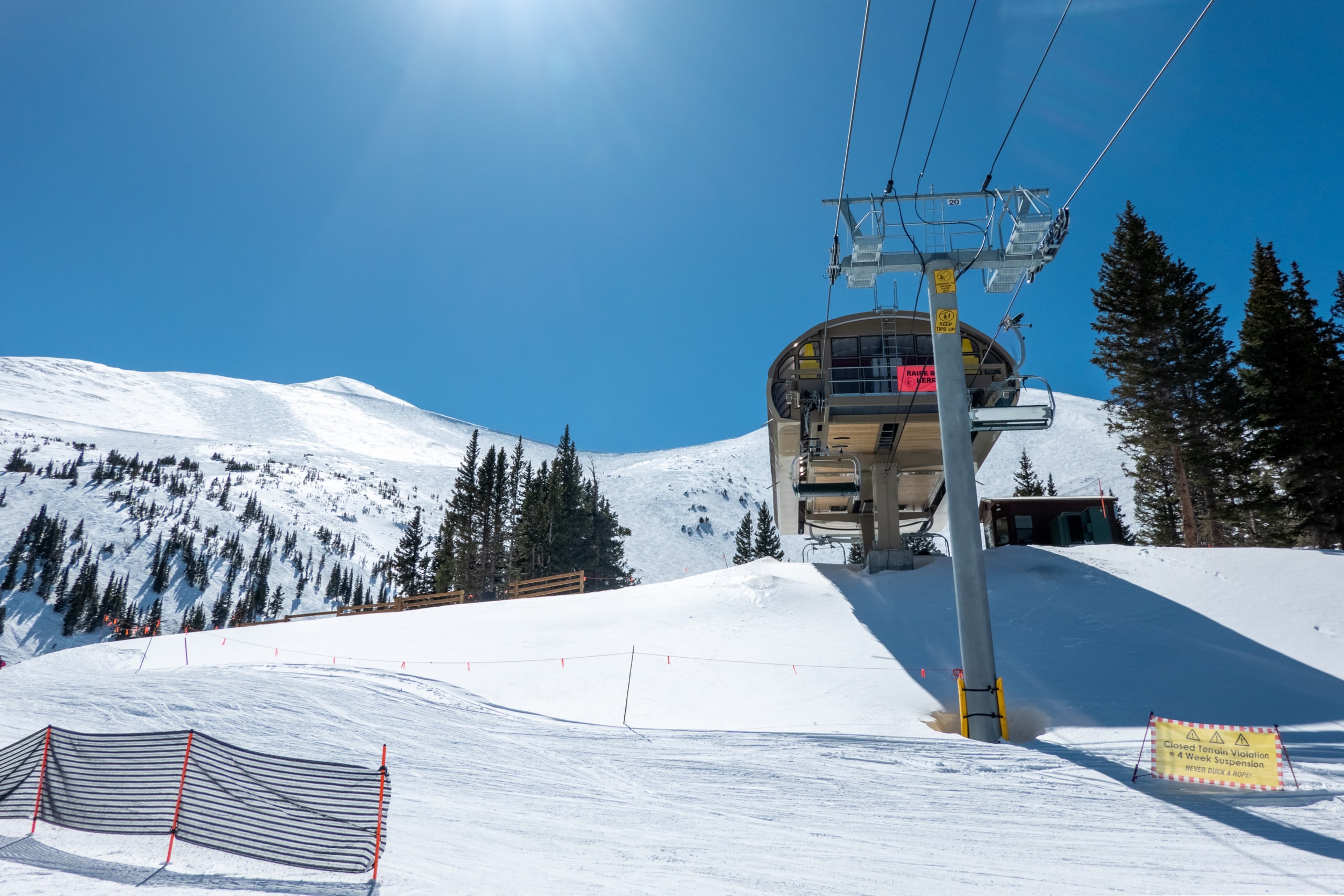 Ski lift rising from a snowy slope under a bright blue sky, with evergreen trees on the hillside nearby.