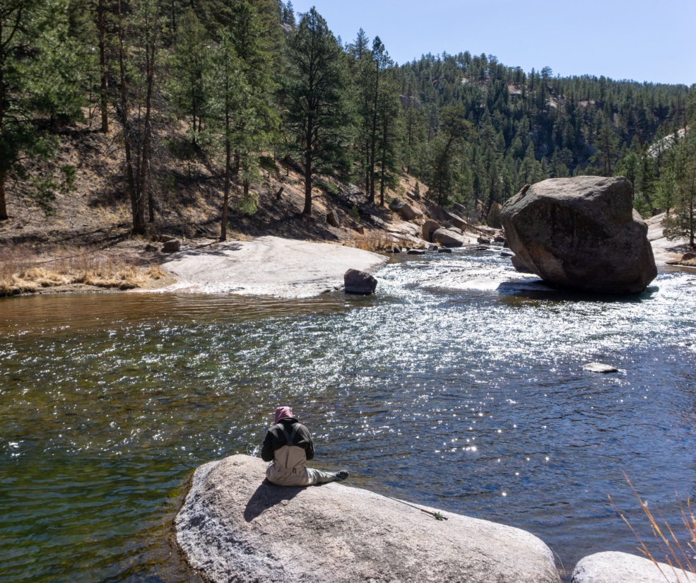 Person sits on a large rock by a sunlit river, forested hills in the background and big boulders along the shore
