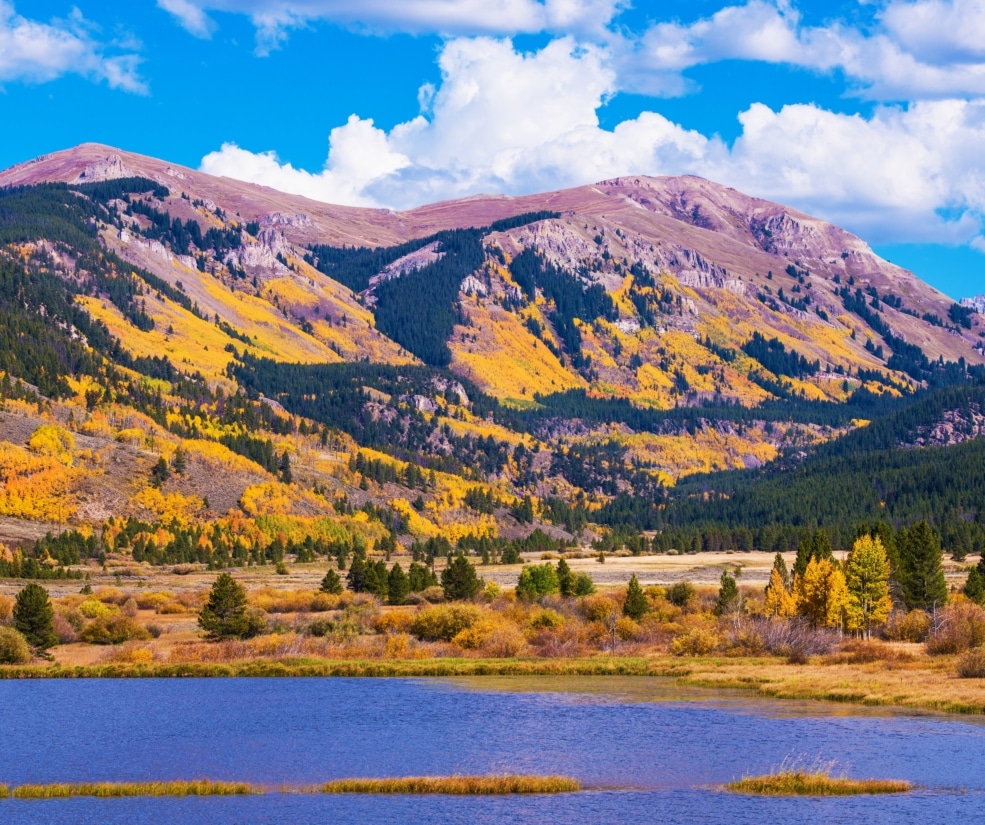 Autumn mountain landscape with yellow and orange trees, evergreen slopes, and a blue lake in the foreground.