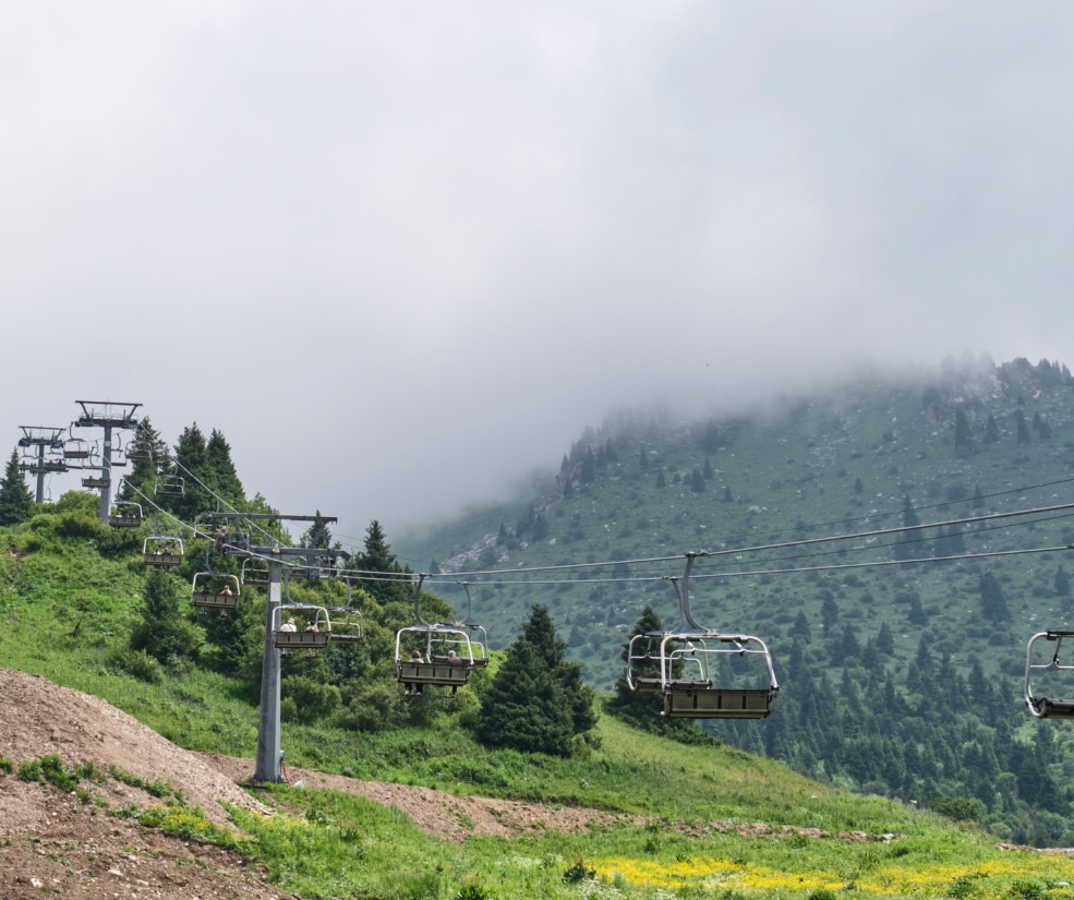 Chairlift with riders glides over a green slope as fog wraps the cloud-covered mountain in the distance.