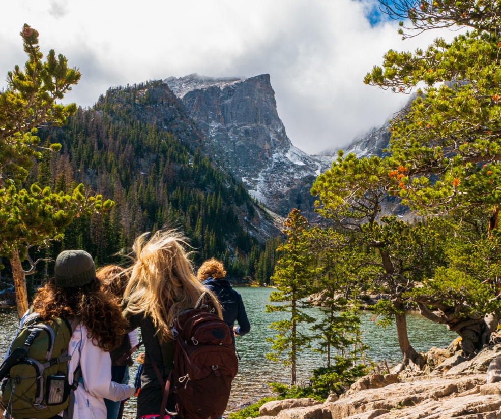 Hikers with backpacks stand at a rocky lakeshore, facing snow-capped mountains framed by pine trees.