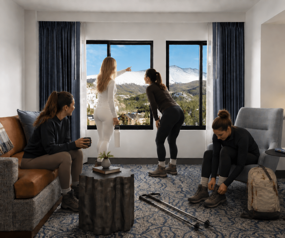 Four women in a cozy hotel room with a mountain view; one points to the window while another leans forward, two others sit and tie laces with hiking gear nearby.