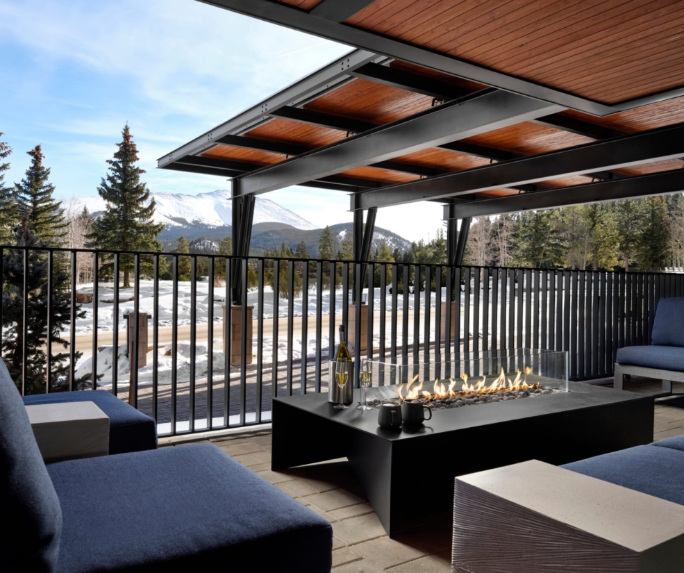 Modern outdoor patio with a rectangular fire pit, blue cushioned seating, and a black railing over a snowy mountain landscape.