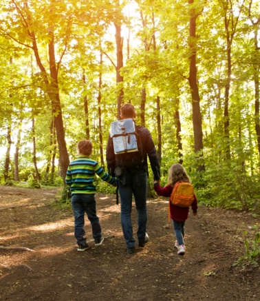 Family of four hiking along a wooded trail with backpacks, sunlight filtering through trees.