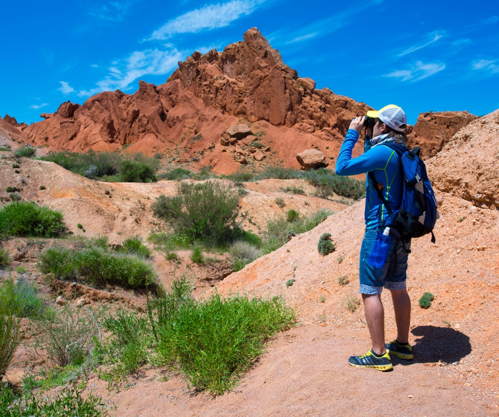 Hiker in a blue jacket with a backpack using binoculars to view the orange-red rock formations under a clear blue sky.